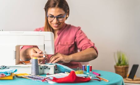 Happy Latin tailor seamstress woman sewing with machine homemade medical face mask for preventing...
