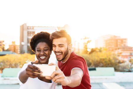Happy couple taking selfie with mobile smartphone outdoor - Mixed race friends having fun with new trendy technology outdoor - Youth millennial generation and multi ethnic teenagers lifestyle conceptの写真素材