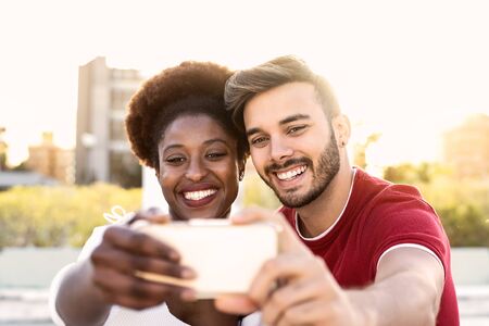 Happy couple taking selfie with mobile smartphone outdoor - Multiracial friends having fun with new trendy social apps - Youth millennial generation and love relationship conceptの写真素材