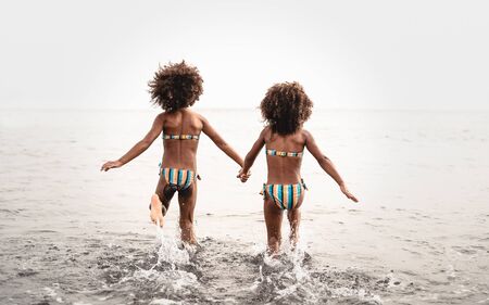 Happy sisters running inside water during summer time - Afro kids having fun playing on the beach - Family love and travel vacation lifestyle conceptの写真素材