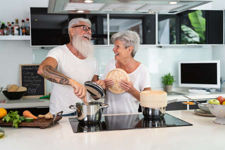 Happy senior couple having fun cooking together at home - Elderly people preparing health lunch in...