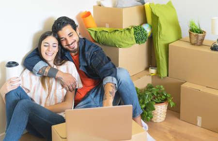 Happy young couple moving in new home first time - Man and woman having fun using computer next...