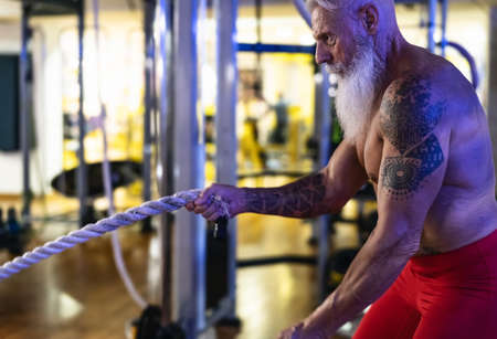 Senior man training with battle rope exercise inside gym - Fit mature male workout in wellness club...