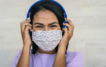 Young woman wearing face mask listening to music with wireless headphones - Latin girl using...