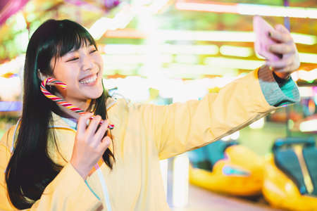 Asian girl taking selfie with mobile phone in amusement park - Happy woman having fun with new...