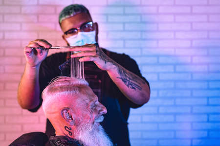Male hairdresser cutting hair to hipster senior client while wearing face surgical mask - Young hairstylist working in barbershop during corona virus outbreak - Health care and haircut salon conceptの写真素材