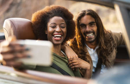 Happy young couple taking selfie with mobile smartphone while doing road trip - Travel people having fun driving in convertible car discovering new places - Relationship and vacation lifestyle conceptの写真素材