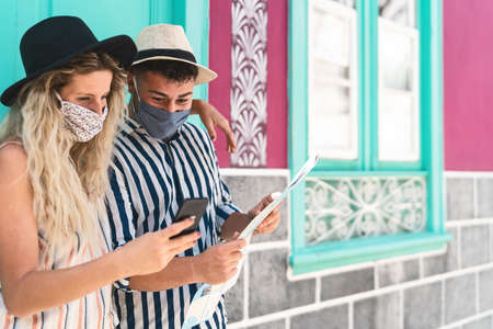 Young couple wearing face protective mask while using mobile phone and holding travel map - Travelers people enjoying first trip after corona virus outbreak - Travel health and technology conceptの写真素材