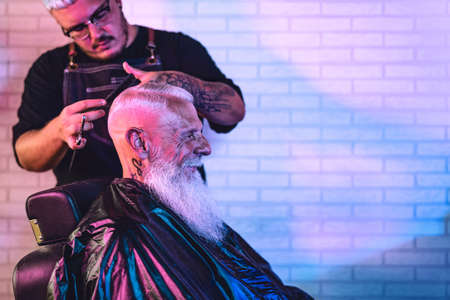Male hairdresser cutting hair to hipster senior client - Side view of young hairstylist working in...