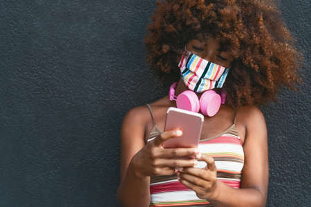 Happy Afro woman using mobile smartphone while wearing face colored mask outdoorの写真素材