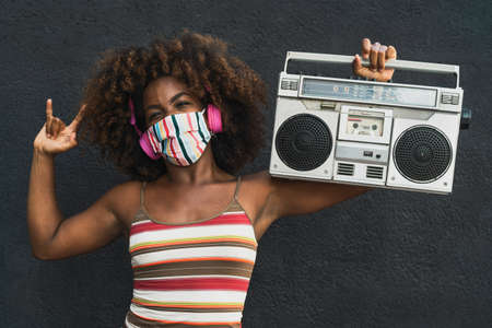 Young Afro woman wearing face mask dancing outdoor while listening to music with wireless headphones...