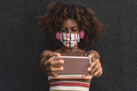 Happy Afro woman taking selfie with mobile smartphone while wearing face colored mask and listening music with wireless headphonesの写真素材
