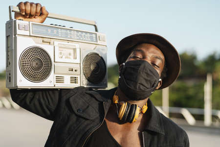 Young African man listening to music with vintage boombox stereo while wearing face mask outdoorの写真素材