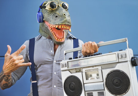 Crazy senior man having fun wearing t-rex mask while listening to music with headphones and vintage boombox stereo outdoorの写真素材