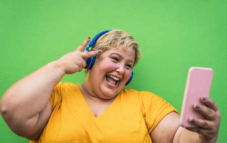 Happy plus size woman taking selfie with mobile smartphone and listening music with wireless headphones - Confident overweight people and technology conceptの写真素材