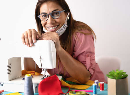 Happy woman working with sewing machine doing homemade protective face mask for preventing and stop...