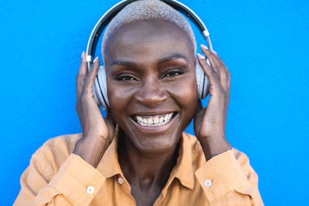 Happy senior African woman having fun listening music with wireless headphones against blue backgroundの写真素材