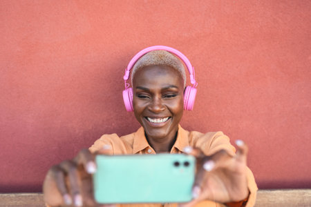 Happy African woman taking selfie with mobile smartphone while listening music with wireless headphonesの写真素材