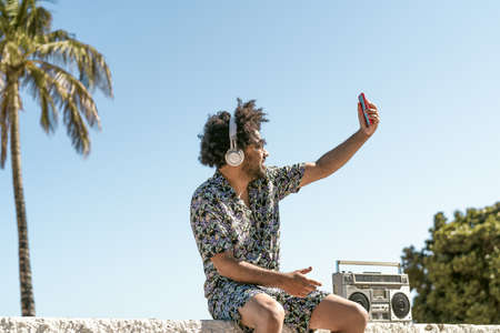 Young man having fun taking selfie with mobile smartphone while listening music with headphones and vintage boombox during summer vacationsの写真素材