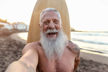 Happy senior surfer taking selfie while having fun surfing at sunset time - Elderly health people lifestyle and extreme sport conceptの写真素材