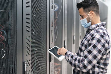 Male informatic engineer working inside server room database while wearing face mask during corona...