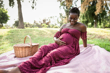 African woman caressing her pregnant belly while doing a picnic in park - Maternity lifestyle...