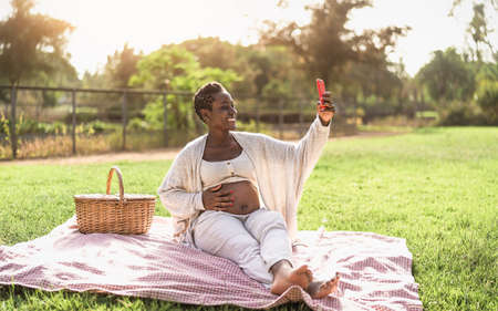 African pregnant woman taking selfie with mobile smartphone while doing a picnic in park - Maternity lifestyle conceptの写真素材