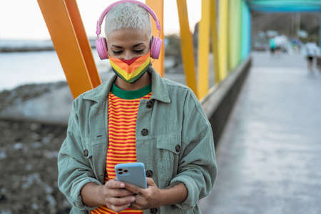 Young gay woman wearing LGBT face mask and using mobile smartphone while listening to music with wireless headphones outdoor - Gender equality and technology conceptの写真素材