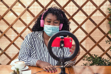 Happy latin woman wearing face mask vlogging online using smartphone cam and ring led led in coffee shopの写真素材