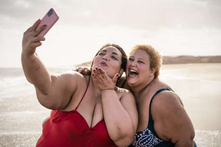 Happy plus size women taking selfie with mobile smartphone on the beach - Overweight friends having fun during vacation timeの写真素材