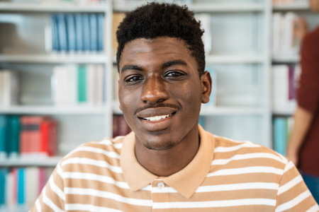 Happy young African man smiling into the camera while standing inside university libraryの写真素材