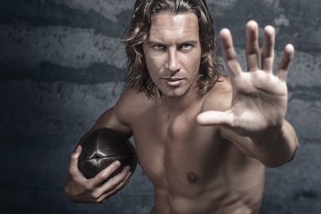 handsome bare chested muscle model is opening his hand in halt gesture and holds football ballの写真素材