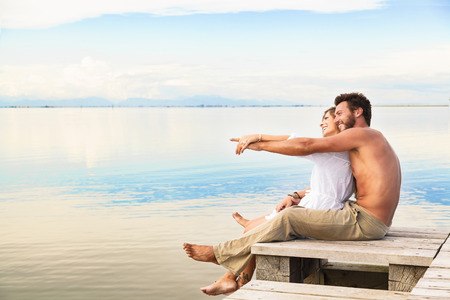 man and woman couple sitting on a jetty and pointing to the horizon under a blue cloudy skyの写真素材