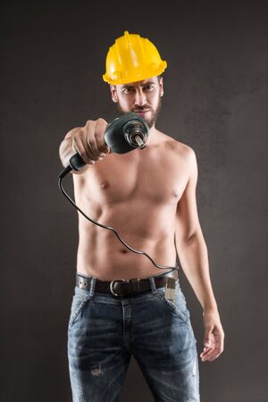 Portrait of attractive bearded workman in jeans and yellow helmet, shirtless, standing and pointing with drill on a gray backgroundの写真素材
