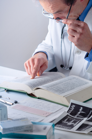 Portrait of Senior Male Doctor looking for Information in a Medical Book at his Office Desktop. Health care, Medical and Pharmacy Concept.の写真素材