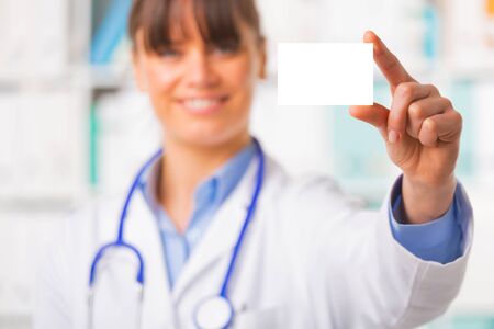 Upper body portrait of young female doctor in white coat with stethoscope, focus on blank business card held between fingersの写真素材
