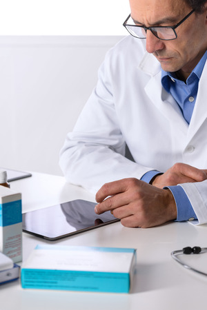 Portrait of Thoughtful Adult male doctor with stethoscope at desk using tabletの写真素材