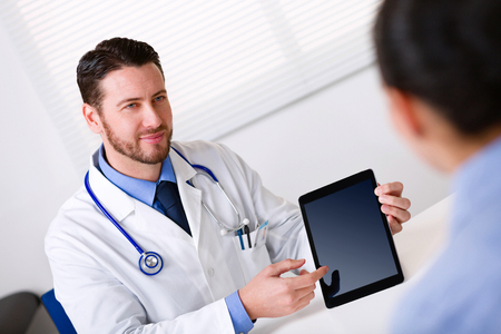Male doctor in white coat showing blank black tablet screen to patient or nurse sitting in front blurred in foregroundの写真素材
