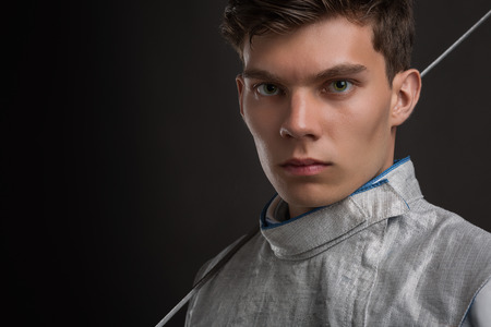 Portrait of Handsome Young male fencer wearing white fencing costume and holding the sword on his shoulders. Dark Background; Looking at cameraの写真素材
