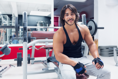 Portrait of young adult Bodybuilder sitting on a bench in a gym and giving the camera a happy friendly smile concept of health and fitnessの写真素材