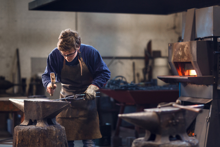 Young blaksmith working with hot metal in a metalworking workshop using specialised tongs and a hammer over an anvil with the furnace in the backgroundの素材 [FY310110668086]