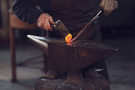 Blaksmith forming a coil with red hot metal from the furnace working with a mallet over an anvil in a close up on the handsの写真素材