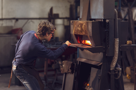 Young blaksmith working in a metalworking workshop holding a set of tongs in a burning furnace as he manufactures a bespoke objectの写真素材
