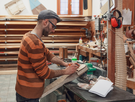 Carpenter man in brown sweater marking wooden detail using pencil, standing at the crafting table in...