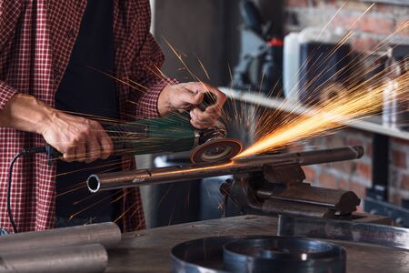 Hands of Metalworker in protective gear working in an engineering workshop sanding a metal bar with an orbital sander with fiery sparksの写真素材