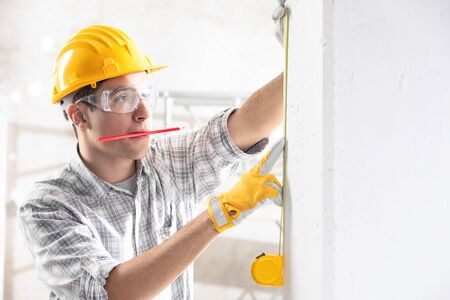 Young construction worker in yellow hardhat, safety goggles and gloves measuring the wall with...