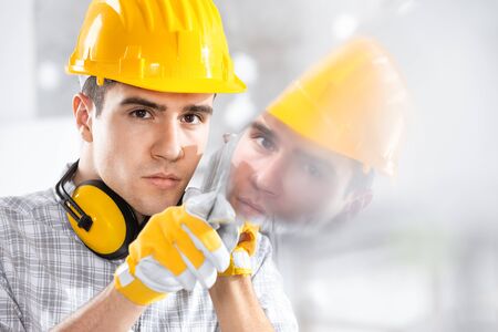 Builder wearing a hardhat and ear muffs reflected in glass he is holding with gloved hands on site
