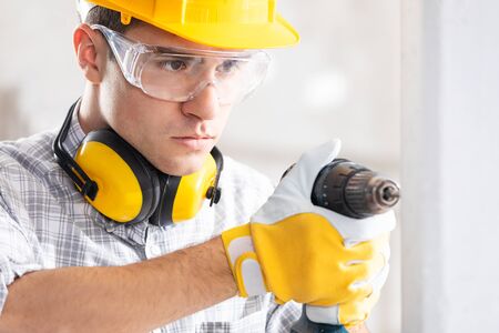 Builder drilling a hole in a new white wall on site with a battery operated hand drill watching what he is doing with a look of concentrationの写真素材