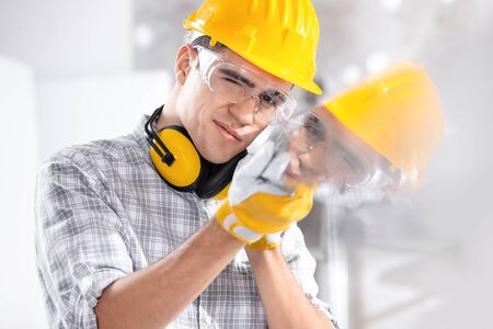 Builder wearing a hardhat and ear muffs reflected in glass he is holding with gloved hands on site