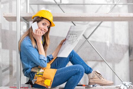 Young female engineer or architect holding a blueprint in her hand as she chats on her smartphone on...